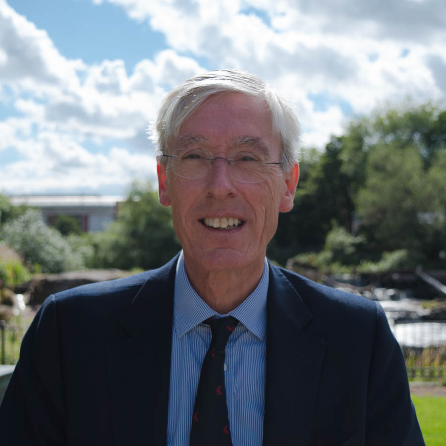 An older man in a suit stands outside, smiling in front of a cloudy sky.